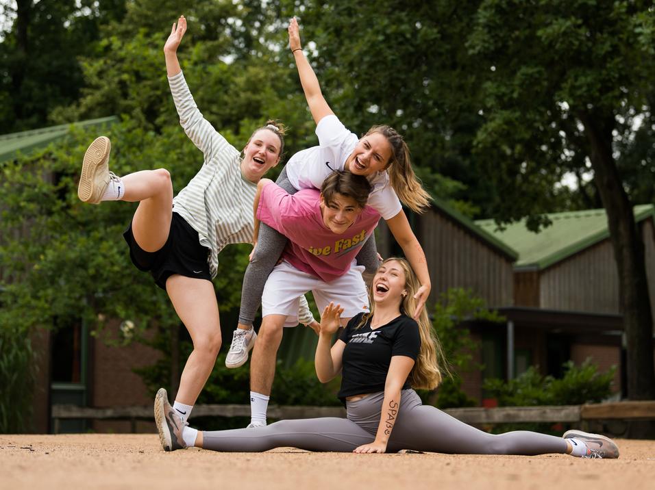 Vier junge Frauen posieren fröhlich auf einer sandigen Fläche, eine sitzt im Spagat, während die anderen sie umarmen.