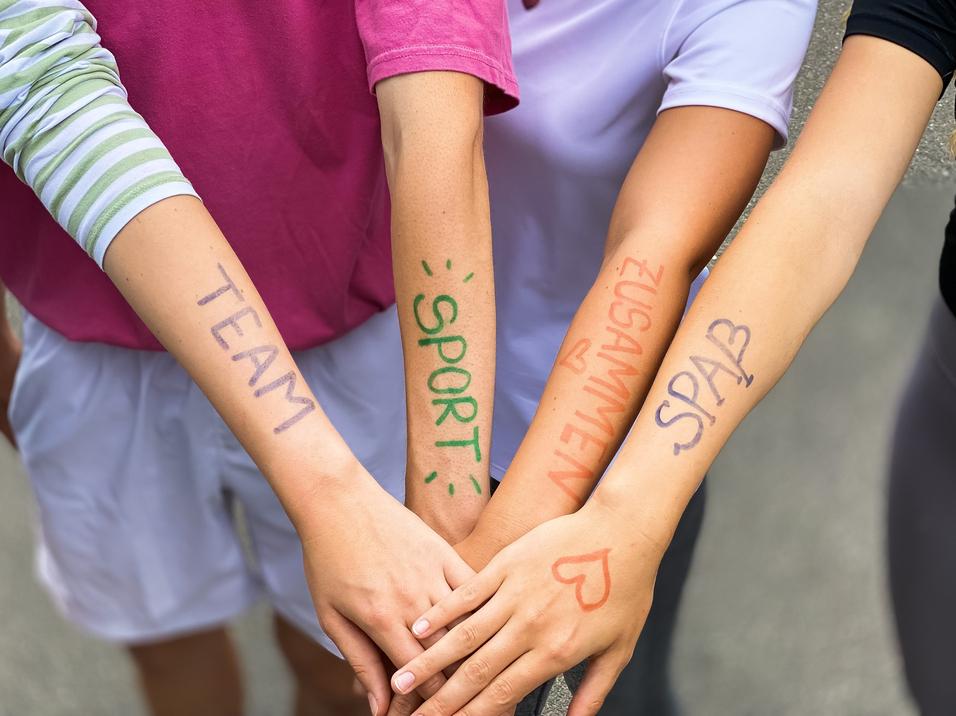 Vier Arme liegen übereinander, mit farbiger Schrift: „TEAM“, „SPORT“, „ZUSAMMEN“, „SPASS“ auf der Haut.