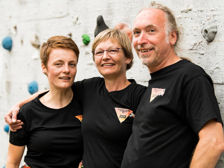 Drei Personen in schwarzen T-Shirts lächeln vor einer Kletterwand mit bunten Griffen.