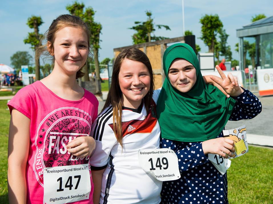 Drei lächelnde Mädchen in Sportkleidung mit Startnummern stehen auf einer Wiese, umgeben von einer Veranstaltung.