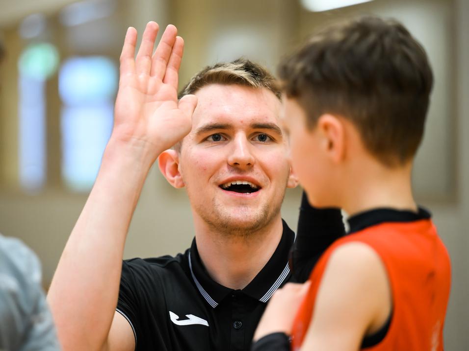 Trainer spricht mit einem jungen Spieler, während er seine Hand hebt, in einer Sporthalle mit Holzwand.