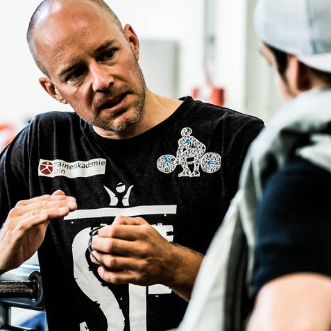 Mann mit glattem Kopf erklärt einem Gesprächspartner etwas in einem Sportstudio.